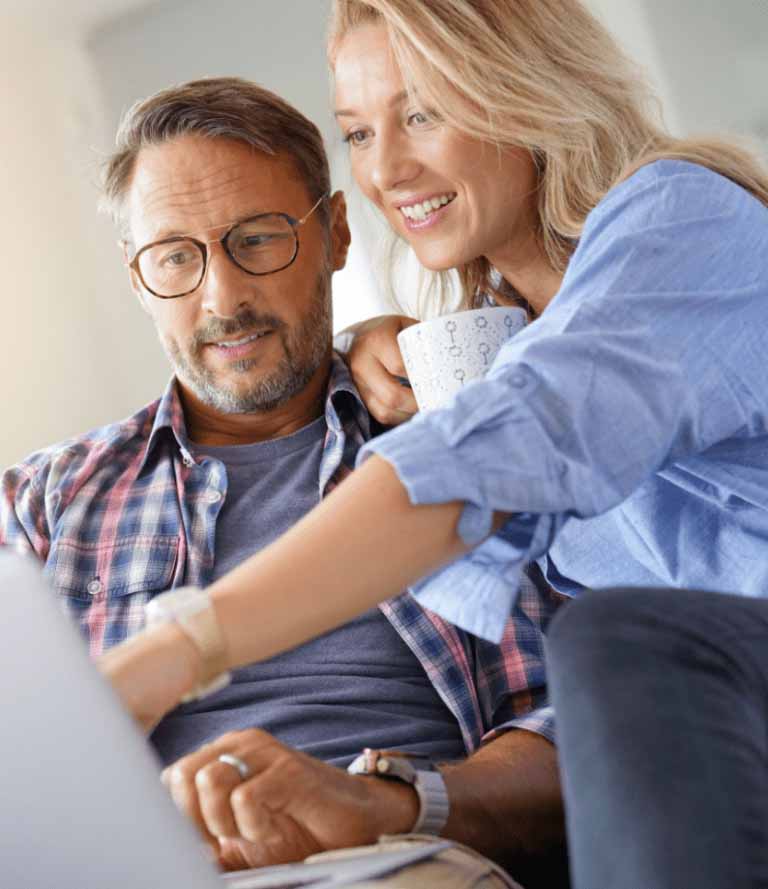 An image of a married couple smiling at their laptop screen. The woman is pointing something out to her husband.